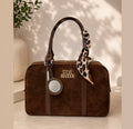 A brown suede handbag with a top handle, a leopard-print scarf, and a circular mirror charm, featuring the 'STEVE MADDEN' logo. The bag is styled on a beige surface with a decorative ceramic vase and plant in the background.