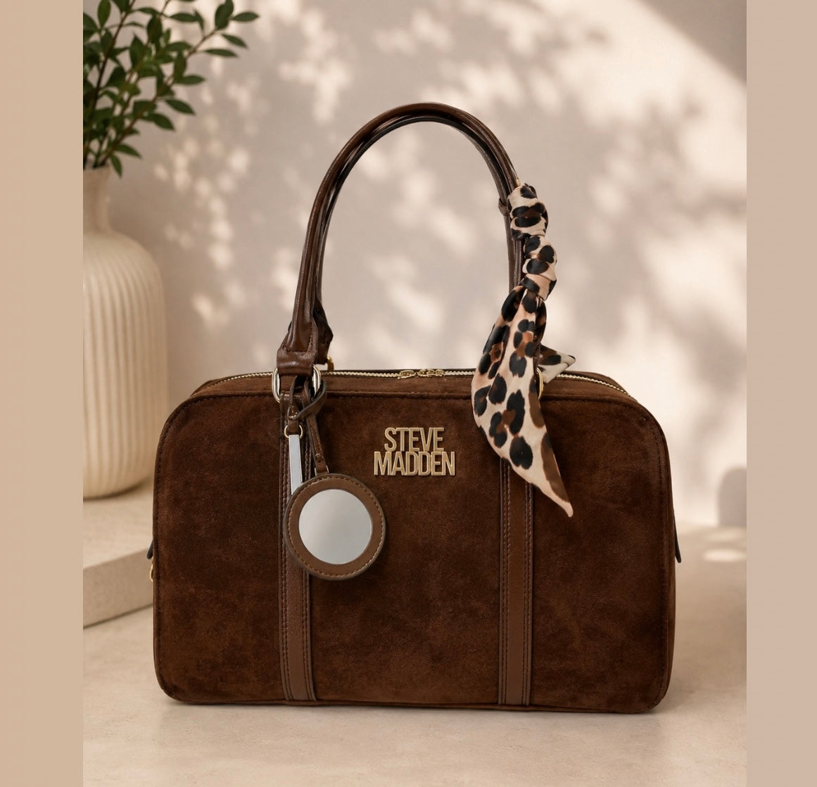 A brown suede handbag with a top handle, a leopard-print scarf, and a circular mirror charm, featuring the 'STEVE MADDEN' logo. The bag is styled on a beige surface with a decorative ceramic vase and plant in the background.