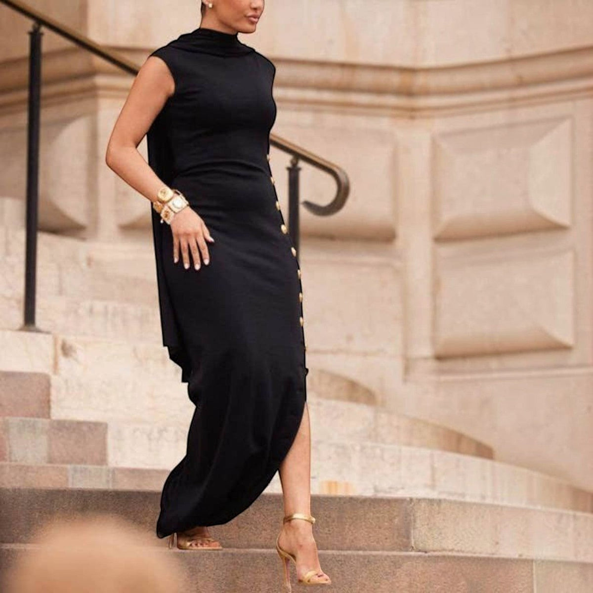 Angie Style Boutique black sleeveless maxi dress with shawl collar, side slit, and gold button details on stone stairs